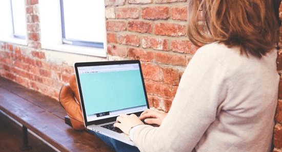 woman sitting on bench with legs up and blogging on laptop beside a brick wall and window